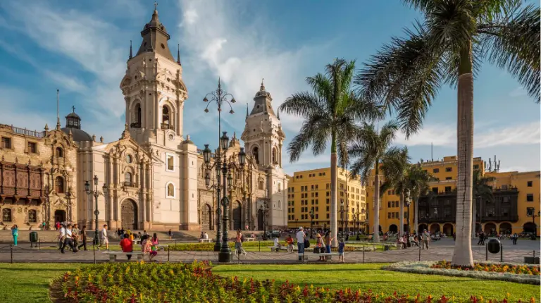 Vista diurna de la Plaza Mayor de Lima, mostrando la Catedral, palmeras y jardines bien cuidados.